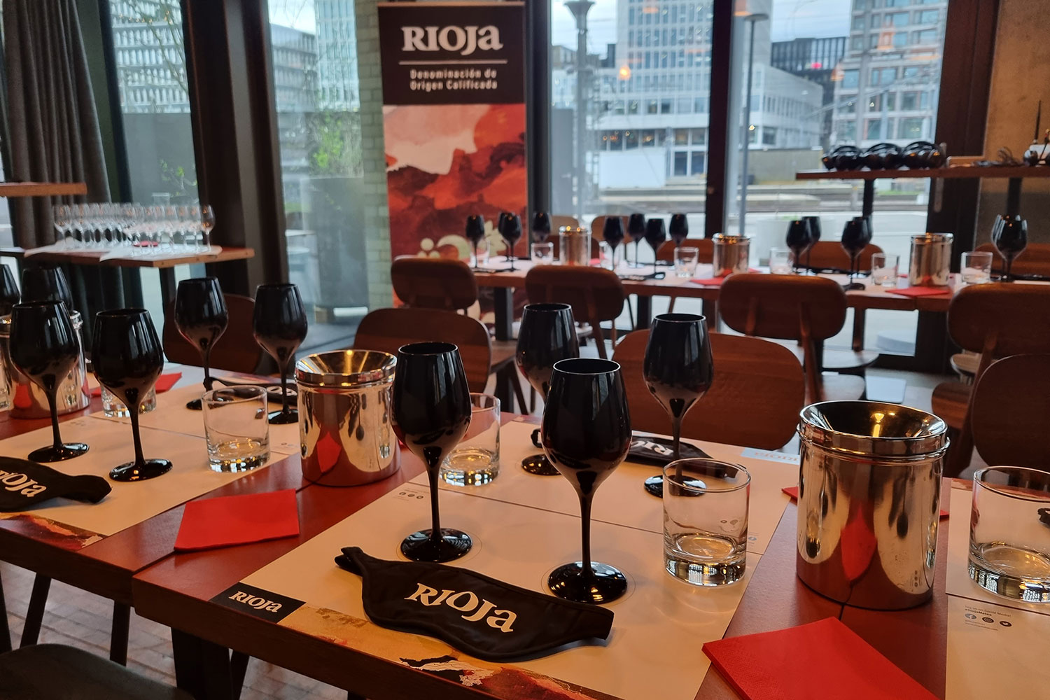 Bild von einer Degustation mit Rioja Branding. Roll-up Banner im Hintergrund, zwei Tischreihen mit Degustationsset: schwarze Gläser und Augenbinde.