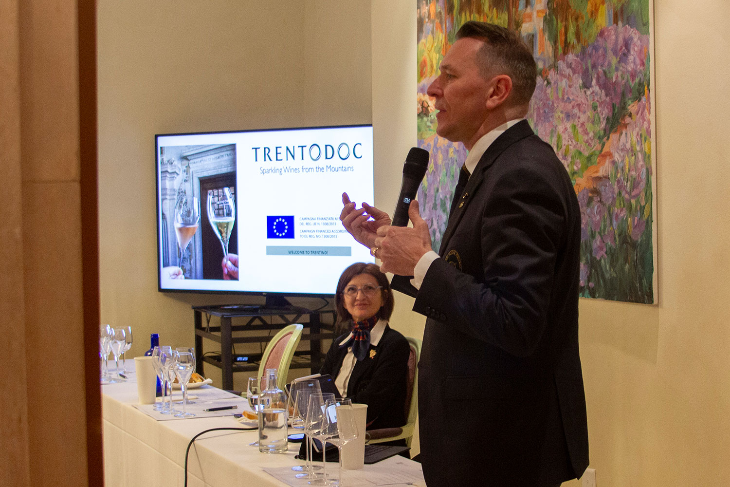 Trentodoc Lugano Roberto Anesi hält ein Mikrofon und spricht bei einer Präsentation, während im Hintergrund eine Leinwand mit der Aufschrift 'Trentodoc' und Bildern von Sektgläsern zu sehen ist. Anna Valli sitzt am Tisch und hört aufmerksam zu. Auf dem Tisch stehen mehrere Weingläser und Wasserflaschen.