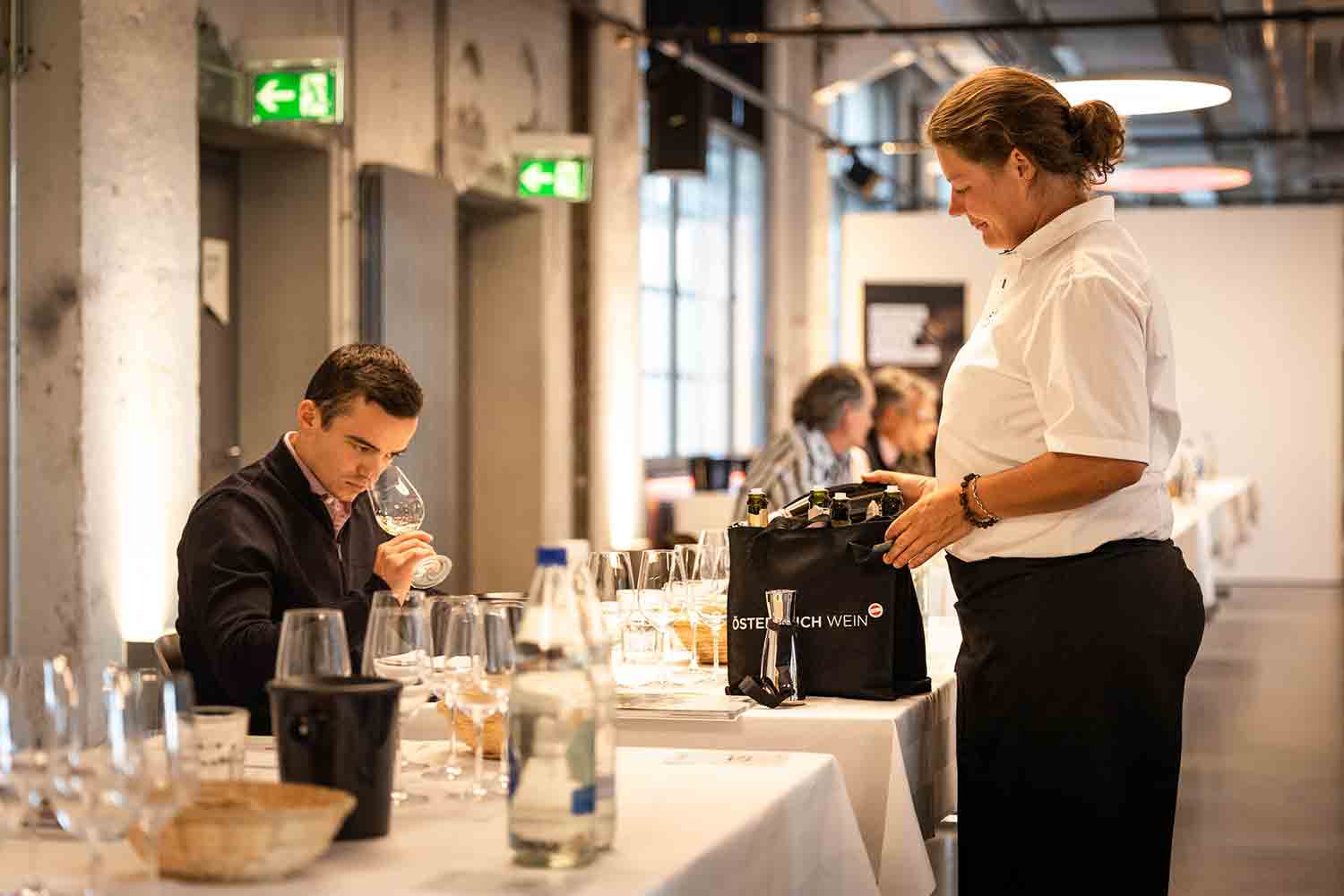 Austrian Flight Tasting 2024 Weindegustation mit einer Person, die sorgfältig ein Glas Wein prüft. Im Hintergrund steht eine Servicemitarbeiterin mit einer Tasche, die mit 'Österreich Wein' beschriftet ist, was auf eine Verkostung österreichischer Weine hinweist. Auf dem Tisch sind mehrere Weingläser und Flaschen angeordnet, was zur Atmosphäre der Degustation beiträgt.