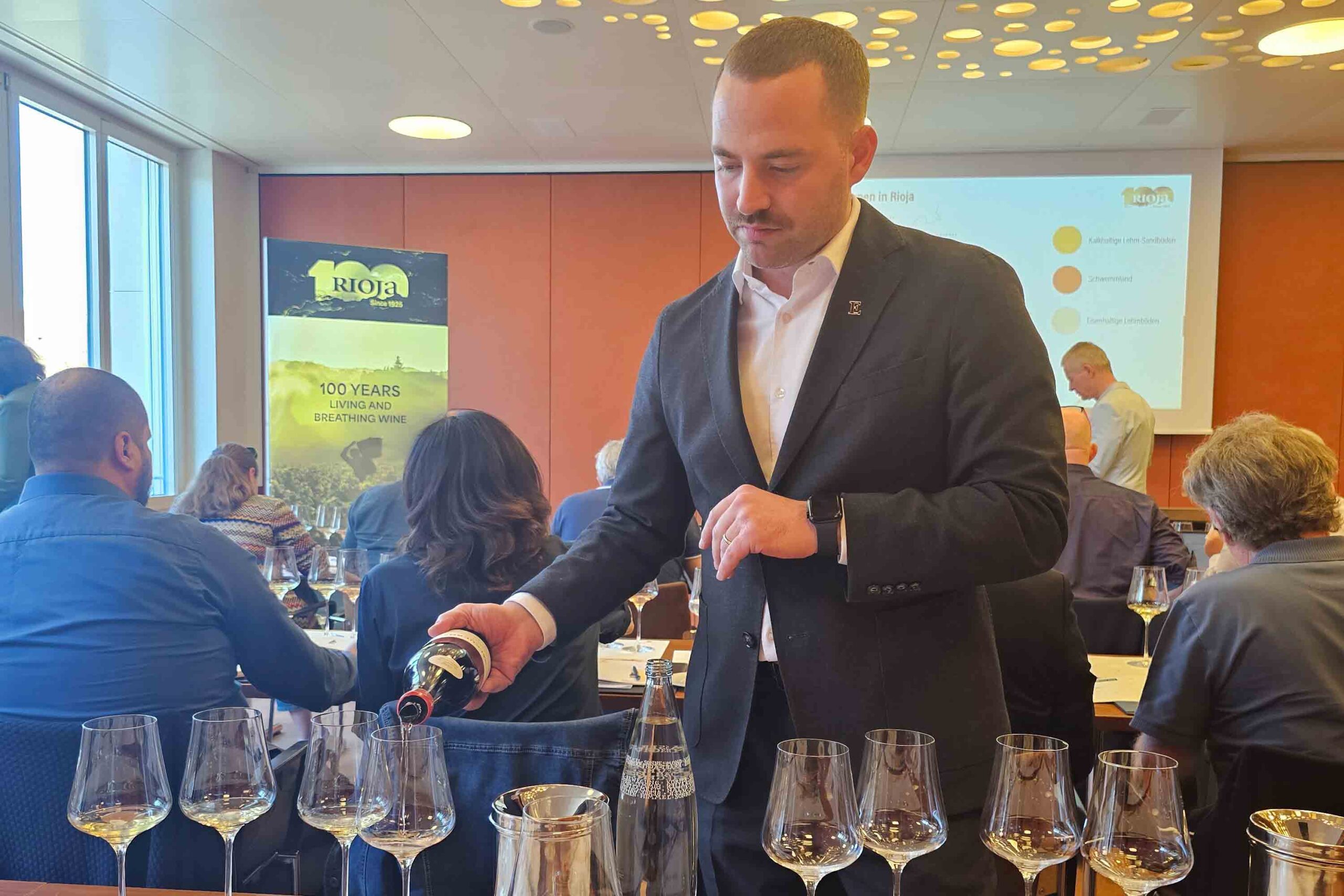 Loris Lenzo, Sommelier, schenkt bei einer Rioja-Weinverkostung im Einstein St. Gallen Weisswein ein. Im Hintergrund ein Banner mit dem Rioja-Logo und dem Slogan „100 Years Living and Breathing Wine“ zum 100-jährigen Jubiläum der Weinregion. Teilnehmer folgen der Präsentation im gut gefüllten Verkostungsraum.
