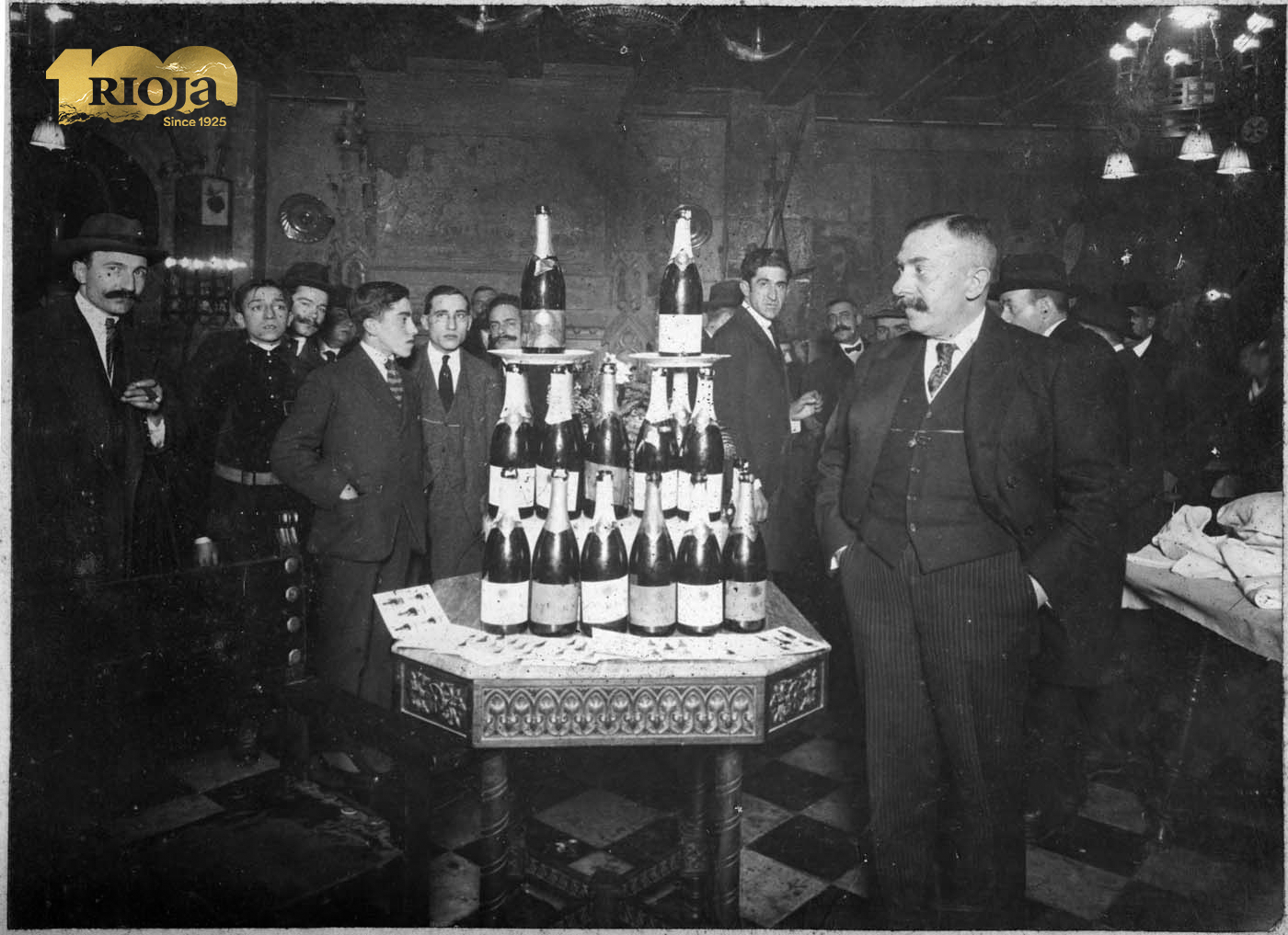 Historisches Schwarz-Weiss-Foto zur Feier von 100 Jahren Rioja: Männer in Anzügen versammeln sich um einen Flaschenturm in festlicher Atmosphäre – Jubiläumsbild zum Rioja-Centenary.