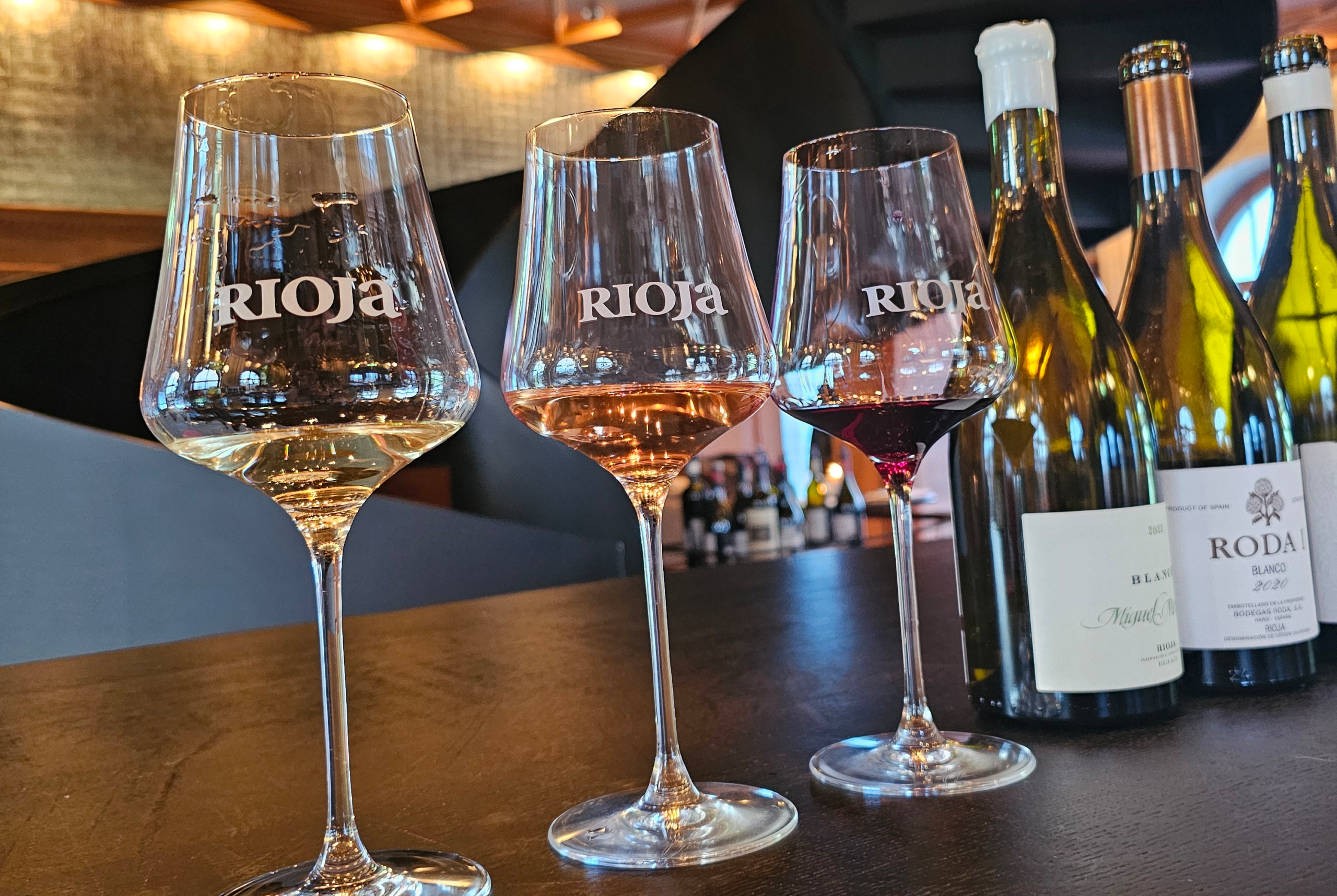 Rioja Prestige Tasting: drei Weingläser mit Weiss-, Rosé- und Rotwein sowie Rioja-Weinflaschen im Hintergrund auf einer Degustationsbar